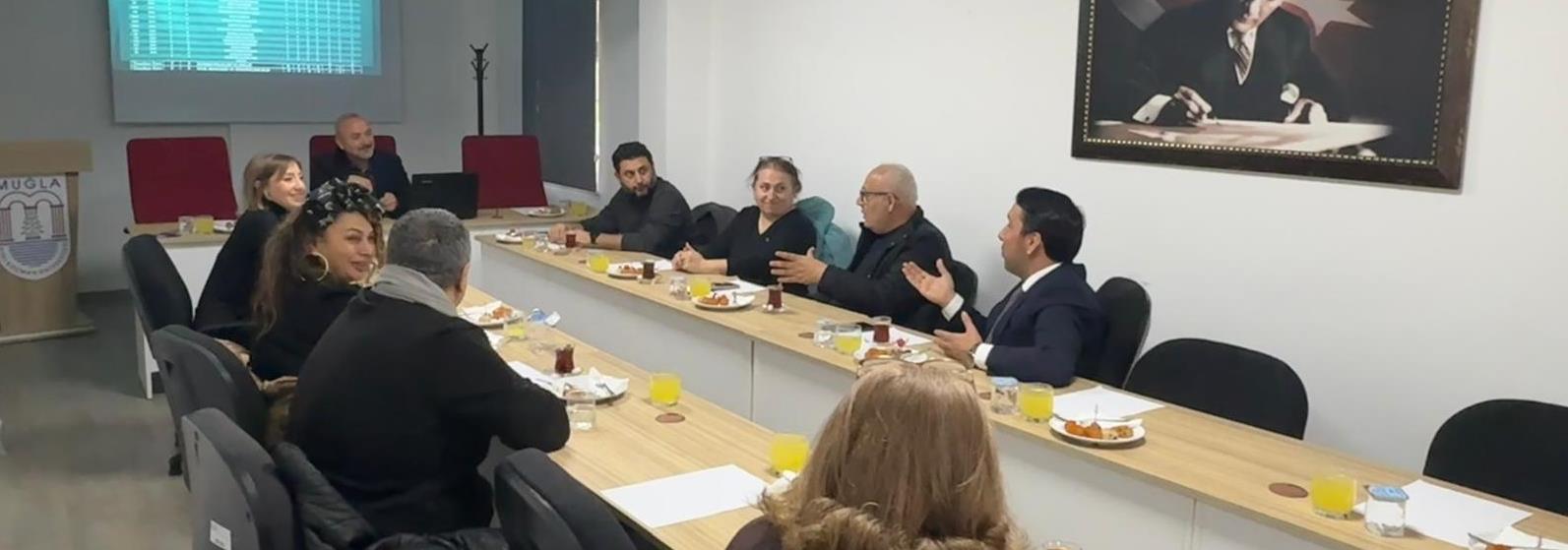 U bir masa etrafında kadın ve erkeklerden oluşan yaklaşık 10 kişi gülümseyerek konuşuyor. 
